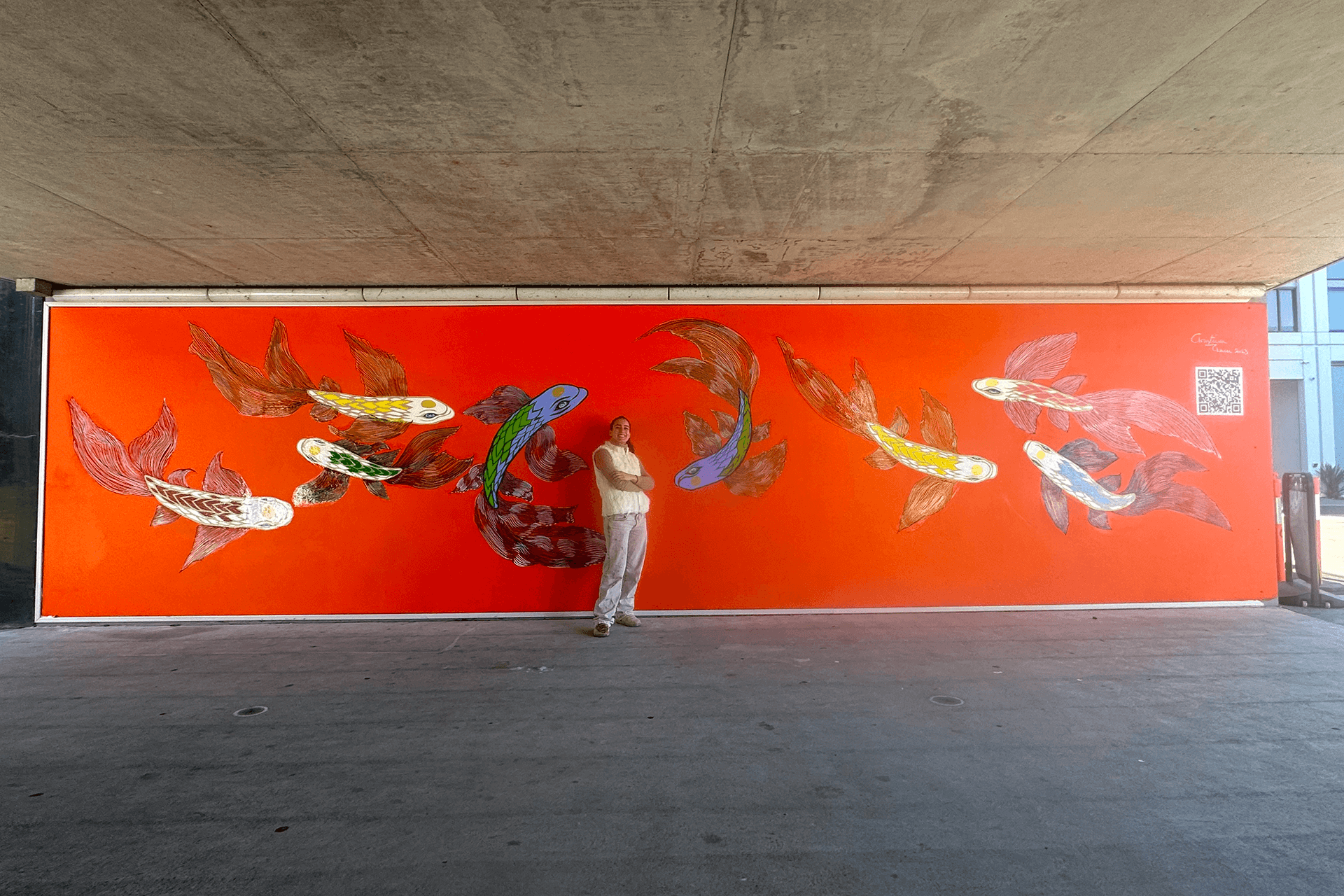 Artist Christina Tsaou, smiling and standing in front of completed mural spanning an entire wall at the Brushbox Street rail underpass, depicting eight koi fish in different colours on orange backdrop, lit by daylight. Image supplied by Artist.